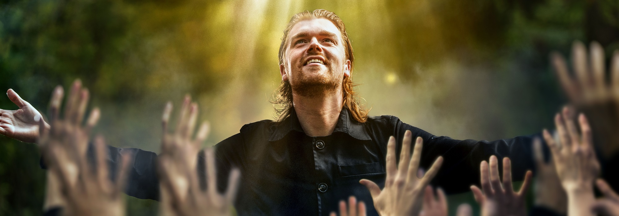 Man met uitgestrekte armen in een bosachtig decor, terwijl zonlicht op hem valt en handen uit het publiek naar hem reiken tijdens de Passiespelen in Tegelen.