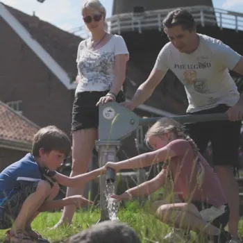 Vader pompt water uit een oude handpomp terwijl de kinderen het water opvangen en de vrouw staat te kijken