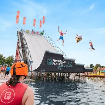 Volwassenen springen van de Super Slide het water in bij Fun Beach Panheel, met toeschouwers en een zomerse sfeer op de achtergrond.